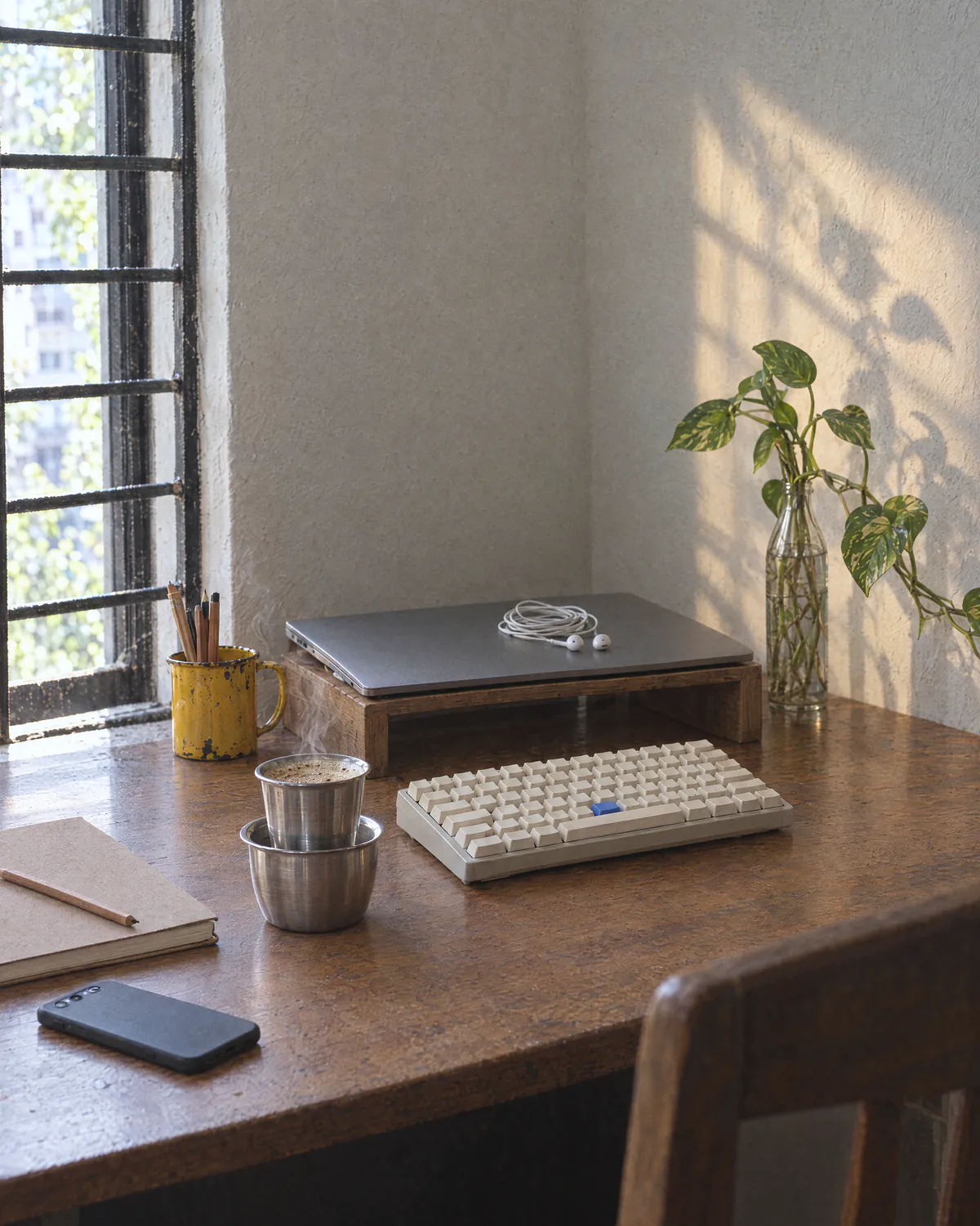 The same desk early in the morning: the laptop closed with earphones on the lid, a steaming steel tumbler of coffee, a yellow mug of pencils, and a plant’s shadow on the wall.